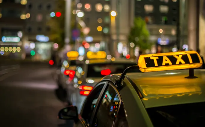 Taxi with a lit roof sign in a busy street at night.
